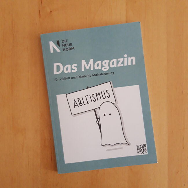 Foto: Die Zeitschrift "Die Neue Norm - Das Magazin ... für Vielfalt und Disability Mainstreaming" liegt auf einem Holztisch. Zu sehen ist der Titel des Magazins mit einer Zeichnung: Ein Gespenst hält ein Schild hoch, darauf steht: Ableismus.